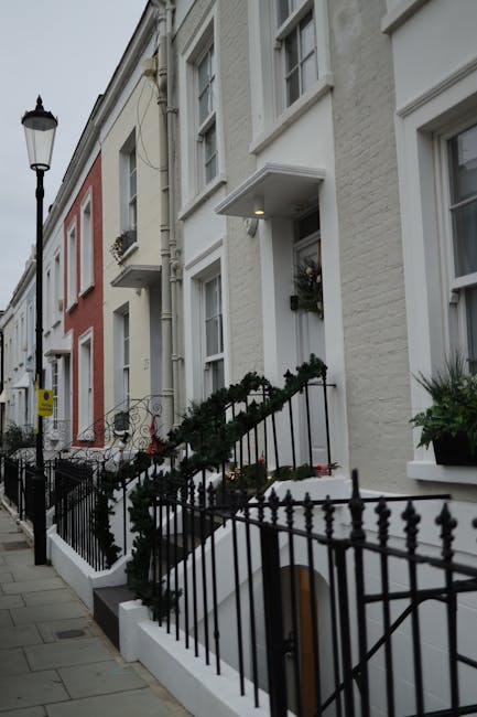 Exterior view of a pink and white terraced house with the number 53 displayed above a vibrant red front door, accessed by a short staircase with black iron railings. A small green shrub is situated ne