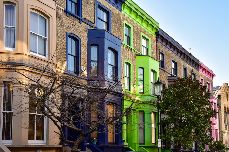 A row of residential buildings in Notting Hill with façades painted in various bright colors, including beige, deep blue, vibrant green, and pink. The buildings feature large, white-framed sash windows, some with curtains visible inside, and decorative architectural details around the windows and rooflines. In front of the buildings, a leafless tree with thin, twisting branches partially obscures the lower windows. A classic black street lamp is situated on the pavement near the green building. The scene is well-lit with natural daylight, and the sky visible at the top of the image is clear and blue. This colorful street scene is representative of the lively neighborhood, often associated with house removals and property relocations in Notting Hill, as depicted by Notting Hill Man and Van, specialists in removals and home relocation services.