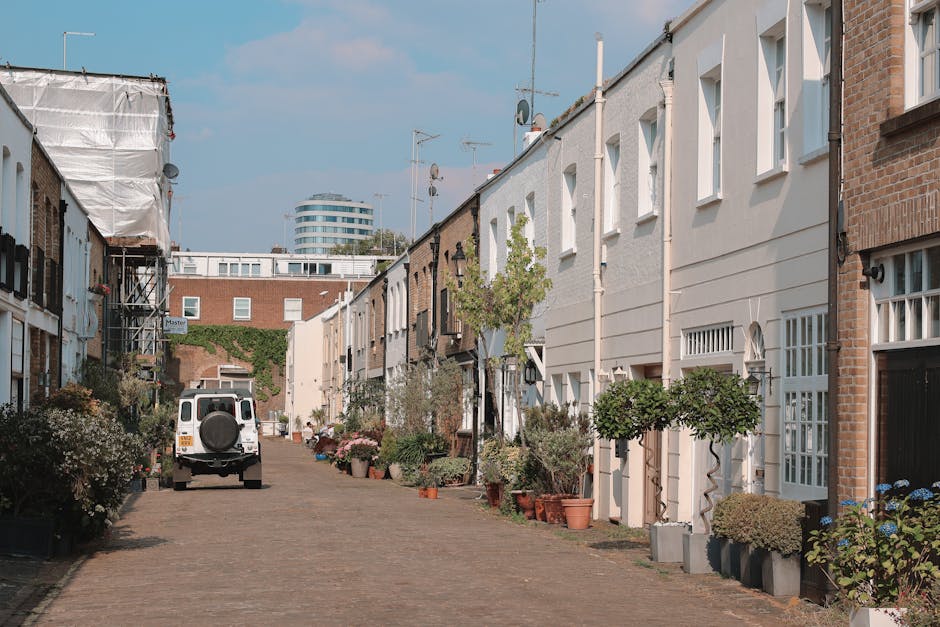 A quiet residential street in Notting Hill with terraced houses featuring white and brick exteriors, some with small front gardens decorated with potted plants and trees. In the foreground, a white van with a spare tire mounted on the back is parked on the cobblestone pavement. Near the van, a person is seen lifting or positioning a large cardboard box wrapped in plastic, possibly during a house removal or moving process. To the right, several furniture pieces and boxes are lined up on the pavement, ready to be loaded into the van or moved into a nearby home. The scene is set during daylight with a clear, partly cloudy sky, and the environment suggests a typical home relocation involving packing, transporting, and unloading household items. Notting Hill Man and Van is involved in the logistics of this home relocation, providing furniture transport and moving services within the area.