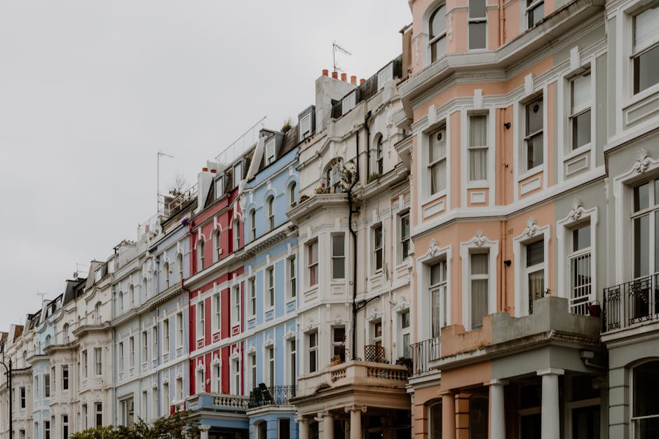 A row of multi-storey terraced houses with Victorian-style architecture, featuring bay windows and decorative mouldings on each façade. The buildings are painted in pastel shades of peach, light blue, and white, with black railings on small balconies and wooden window frames. The upper floors have pitched roofs with chimneys, and some rooftop antennas are visible. The sky is overcast, casting diffused light across the scene. This residential street is located in a typical London neighbourhood, with the lower parts of the buildings opening onto pavements. The image captures the exterior of the houses, emphasizing their architectural details and vibrant colours. This setting is relevant for house removals, illustrating the typical environment where furniture transport and home relocation services by Notting Hill Man and Van might be performed, especially during furniture loading, packing, or moving logistics inside such properties.