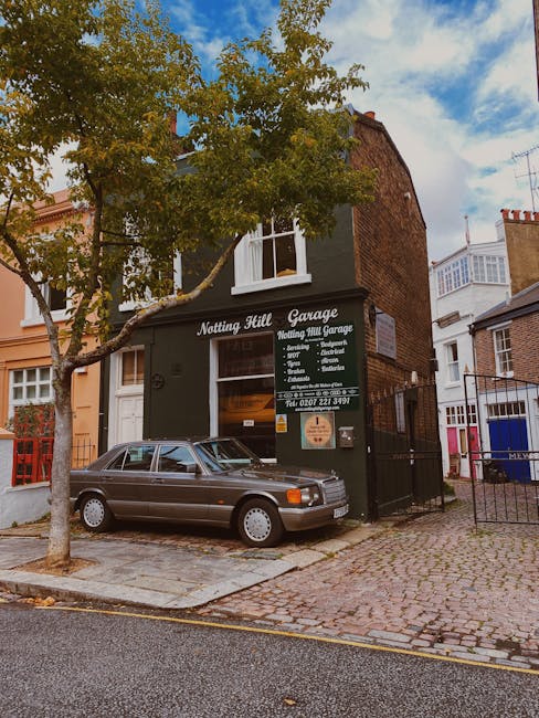 A grey classic car parked on a cobbled street in front of a dark green building with a sign reading 'Notting Hill Garage' that lists services such as removals, garage services, and packing. The garage is part of a two-storey building with a white-framed window on the upper floor and a glass door entrance on the ground level. To the left, a tree with green leaves partially obscures the garage exterior. Inside the garage, visible through the glass door, are packed cardboard boxes and packing materials, indicating preparation for a home relocation or furniture transport. The surrounding area features other residential buildings, with some brick façades and balconies, suggesting a typical London neighbourhood. The scene is captured during daylight with a partly cloudy sky, emphasizing the outdoor loading process in a professional moving context, as conducted by Notting Hill Man and Van.