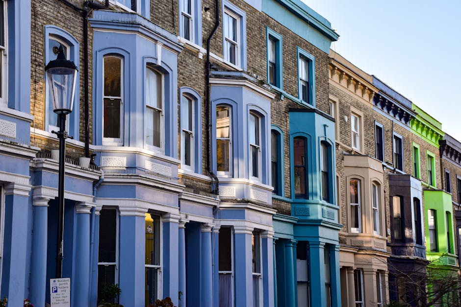 A row of Victorian terraced houses in Westbourne Grove, Notting Hill, with facades painted in pastel shades including light blue, beige, and bright green. The buildings feature large sash windows framed with decorative mouldings and some have small balconies or bay windows. A black vintage-style street lamp is positioned on the pavement in front of the houses. The scene shows part of a home relocation process with outdoor furniture, cardboard boxes, and plastic wrapping visible on the sidewalk, indicating packing and moving preparations. A Notting Hill Man and Van van is parked on the street, partially visible, ready for furniture transport and loading. The overall environment appears sunny with clear skies, emphasizing the residential and historic character of the area, suitable for professional removals and house moving services.