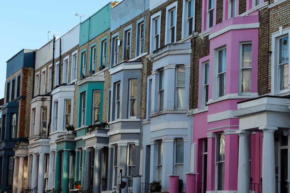 An exterior view of a pink, curved residential building with multiple white-framed windows set in a vertical arrangement, located on a street with a black iron fence along the sidewalk. The building's
