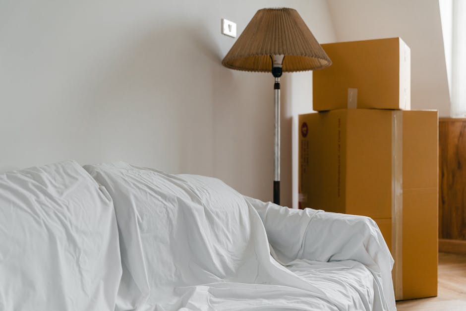 Image showing a packing and moving scene inside a room with neutral-colored walls and wooden flooring. Several cardboard boxes of various sizes are stacked near a wooden cabinet, with some boxes sealed with packing tape and others open, revealing packing materials. A floor-standing lamp with a brown shade is turned on, casting ambient light across the room. In the foreground, there is a white sofa with a slightly wrinkled slipcover. The sofa is positioned adjacent to the boxes, suggesting a home relocation process involving packing, furniture disassembly, and transport preparations, with the setting indicating an organized and tidy living space before or during the move. Notting Hill Man and Van specializes in furniture transport and house removals, supporting efficient loading and transportation services.