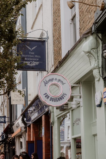 Photograph of a narrow street in Notting Hill with several business signs hanging above shop fronts. The image features a black sign for 'The Notting Hill Bookshop' and a pink illuminated sign shaped like a doughnut with the text 'Always a good FOR DOUGHNUTS' written on it. The buildings are constructed from brick and painted wood, and there are pedestrians partially visible at the bottom of the frame. The scene captures an outdoor urban environment during daytime, with diffused natural lighting. While no furniture or moving equipment is visible, the image is relevant for a house removals service context focused on transportation logistics in residential or commercial areas, illustrating typical local surroundings where furniture and household items might be moved or delivered.
