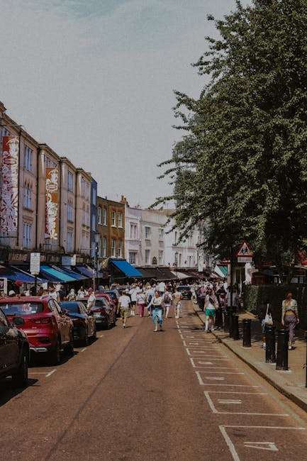 A busy street scene in Portobello Road, with pedestrians walking along the pavement and browsing market stalls under blue awnings on the left side of the image. Parked cars line both sides of the narrow road, which features a designated loading zone marked with white dashed lines and numbers. In the background, rows of colorful terraced townhouses with distinctive facades and large windows extend along the street. A large leafy tree on the right provides partial shade over the scene. The lighting suggests a sunny day, with clear skies overhead. The image captures the typical vibrant atmosphere of Portobello Market on market days, with residents and visitors engaging in shopping and strolling. Notting Hill Man and Van, known for their home relocation and furniture transport services, often operate within such busy environments, managing packing, loading, and moving logistics amidst the bustling street scene.