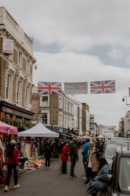 A busy street scene on Portobello Road in Notting Hill, lined with market stalls covered by colorful awnings and a white canopy, with people browsing and shopping. Some individuals carry shopping bags or stand next to tables displaying various goods, while others walk or sit on benches along the pavement. The street is filled with pedestrians, including men and women dressed in casual attire, some engaged in conversation. Vehicles, including a grey van, are parked along the curb or being loaded with household furniture and boxes wrapped in plastic or cardboard. Overhead, banners with the Union Jack flag and a sign reading 'PORTOBELLO ROAD W11' are strung across the street, indicating the market's location. The surroundings include multi-story buildings with shopfronts, one of which has a sign for a local business, and the natural daylight illuminates the scene, capturing the essence of a vibrant market day aligned with house removals and moving services.