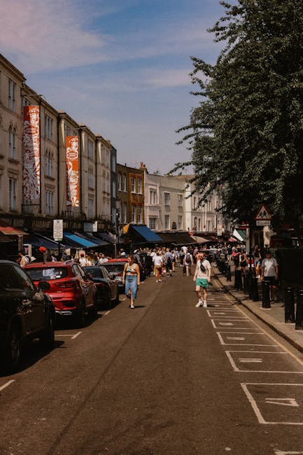A busy street scene in Portobello Road, with pedestrians walking along the pavement and browsing market stalls under blue awnings on the left side of the image. Parked cars line both sides of the narrow road, which features a designated loading zone marked with white dashed lines and numbers. In the background, rows of colorful terraced townhouses with distinctive facades and large windows extend along the street. A large leafy tree on the right provides partial shade over the scene. The lighting suggests a sunny day, with clear skies overhead. The image captures the typical vibrant atmosphere of Portobello Market on market days, with residents and visitors engaging in shopping and strolling. Notting Hill Man and Van, known for their home relocation and furniture transport services, often operate within such busy environments, managing packing, loading, and moving logistics amidst the bustling street scene.