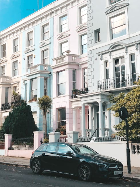 A black hatchback car parked on a residential street in front of a row of Victorian-style terraced houses painted in pastel shades of white, pink, and blue, with decorative iron balconies and large sash windows. The pavement includes a small garden area with shrubbery and a tall palm-like tree. The image captures the exterior environment during daylight with bright, clear weather. The scene reflects typical urban home relocation surroundings, possibly related to house removals or moving services provided by Notting Hill Man and Van, with visible elements such as the parked vehicle and residential facades indicating a setting for furniture transport and moving logistics.