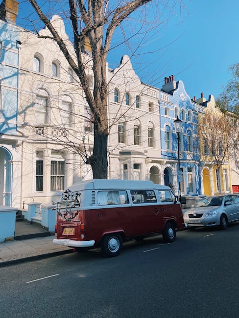 A classic Volkswagen Type 2 van painted in red with a white roof, parked on a residential street in front of a row of colorful Victorian-style townhouses. The van is equipped with roof racks carrying various items, and a bicycle mounted on the rear. Nearby, several cardboard boxes and packing materials are placed on the sidewalk, indicating a house relocation process. The street is lined with leafless trees and the weather is clear with bright blue skies. The scene appears to be part of a moving or packing operation, as often handled by Notting Hill Man and Van, with the van positioned close to the property's entrance to facilitate furniture and item transport during packing and loading for a move to Holland Park Mansions.