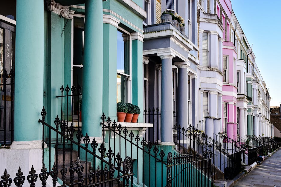 View of a row of terraced Victorian houses in W11 Notting Hill with colorful facades painted in pastel shades of teal, purple, pink, and mint green. The houses feature large windows with white trims, some with flower boxes containing green plants. Each property has black wrought iron railings and gates, with steps leading up to the entrance doors. The buildings have decorative columns supporting small porches, and some have potted plants placed on window ledges. The sidewalk in front is paved, and a clear blue sky is visible overhead, indicating daytime. The scene offers a glimpse into the typical historic architecture in Notting Hill suitable for professional removals and furniture transport during home relocation projects, with [COMPANY_NAME] often providing services in these areas to facilitate smooth packing and moving processes.