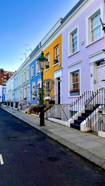 A row of vibrantly painted Victorian terraced houses along a quiet street in Notting Hill during daylight. The houses feature pastel shades, including blue, yellow, pink, and purple, each with white-framed sash windows and small front gardens enclosed by black wrought iron fences. A traditional black street lantern with a green top is mounted on a post in front of one of the houses, casting ambient light. The pavement is clean and paved with concrete slabs, while the asphalt road runs alongside. The houses have external steps leading to front doors, some with black railings, and in the distance, a few houses and urban elements such as television antennas are visible. This scene is associated with house removals and home relocation services, typical of the neighbourhood's characteristic architecture, as offered by Notting Hill Man and Van, highlighting the process of packing, loading, and transporting belongings during a move.