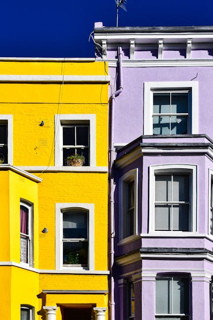 View of a row of terraced Victorian houses in W11 Notting Hill with colorful facades painted in pastel shades of teal, purple, pink, and mint green. The houses feature large windows with white trims, some with flower boxes containing green plants. Each property has black wrought iron railings and gates, with steps leading up to the entrance doors. The buildings have decorative columns supporting small porches, and some have potted plants placed on window ledges. The sidewalk in front is paved, and a clear blue sky is visible overhead, indicating daytime. The scene offers a glimpse into the typical historic architecture in Notting Hill suitable for professional removals and furniture transport during home relocation projects, with [COMPANY_NAME] often providing services in these areas to facilitate smooth packing and moving processes.