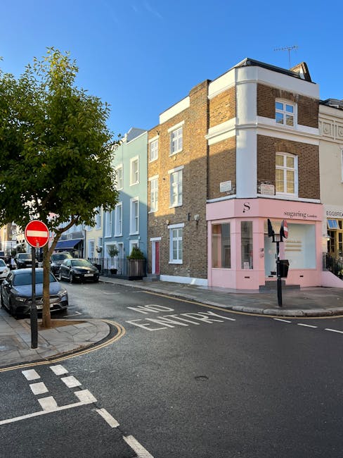 A view of a residential street corner in Notting Hill during daytime, featuring a three-storey building with a ground-floor shop painted in soft pink, displaying large windows with display items inside. Adjacent are other multi-storey buildings with brick and pastel facades, some with white window frames. Parked cars line the curb on the left side of the street, and a tree with green foliage is situated near the corner. The road has clear traffic markings, including a 'NO ENTRY' sign and a 'KEEP LEFT' sign painted on the asphalt, indicating a one-way street. The scene is well-lit with natural sunlight, and the overall environment appears calm and suitable for home relocation or furniture transport activities, with the presence of vans or moving equipment implied by the context of house removals services provided by Notting Hill Man and Van.