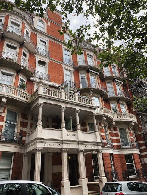 Photograph of a multi-storey red-brick residential building with white stone accents and decorative balconies, viewed from a street lined with trees casting green leaves across the scene. In front of the building, a white van is parked on the pavement, while other vehicles are visible nearby. The building's entrance features a classical-style portico with tall, fluted columns supporting a balcony above, and the address '13-36' etched on the facade. This scene depicts the exterior of a typical London apartment complex often involved in home relocation or removals. Notting Hill Man and Van specializes in house removals, and such a property may be a typical site for furniture transport or packing and moving services, aligned with efficient loading and transport logistics within the urban environment.