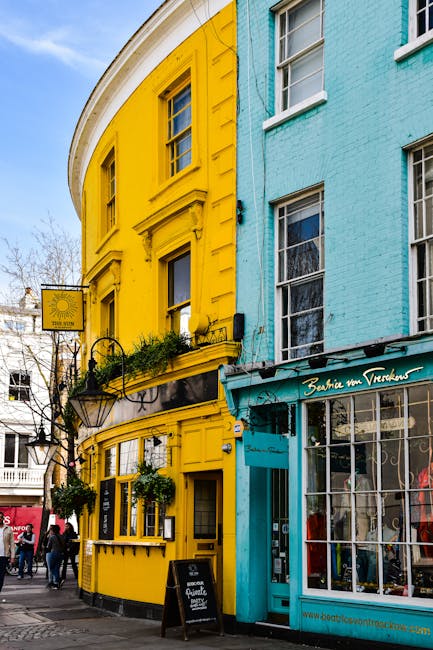 The image shows a colorful street scene outside a corner building with a bright yellow façade on the left and a light blue façade on the right. The yellow section features three rectangular windows, with the middle window having window boxes filled with greenery and plants. Below the windows, there is a black awning with hanging flower baskets and a small chalkboard sign on the pavement. The blue section includes large glass windows with white framing and a sign reading 'Beatrice the Voodooess' above the entrance. On the street, people are walking, and part of a black vehicle is visible, indicating active loading or moving activity. The scene is well-lit with natural daylight, highlighting the vibrant colours. The setting suggests an urban environment suitable for house removals, unloading, and furniture transport, as associated with the services provided by Notting Hill Man and Van.