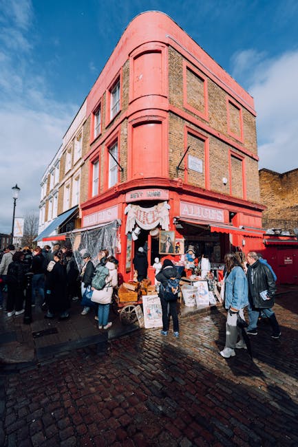 A large, multi-storey building with a curved corner façade painted in shades of pink and red, situated on a cobblestone street. The ground floor features open shop displays with various items for sale, and a canopy with decorative white drapes extends over the storefront. Several people are gathered outside, some browsing, others waiting in line or walking past. The scene is set during daytime with a partly cloudy sky, and the background includes other buildings and a hill. The image illustrates a busy street in Notting Hill, possibly during a market day, with a focus on the architecture, street activity, and retail environment, relevant to house removals and moving logistics as produced or referenced by Notting Hill Man and Van.